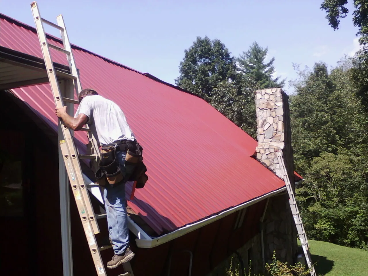 Roofer on a ladder beside a red metal roof and stone chimney — Soffit & Fascia Repair by Roofing Brothers in Molalla