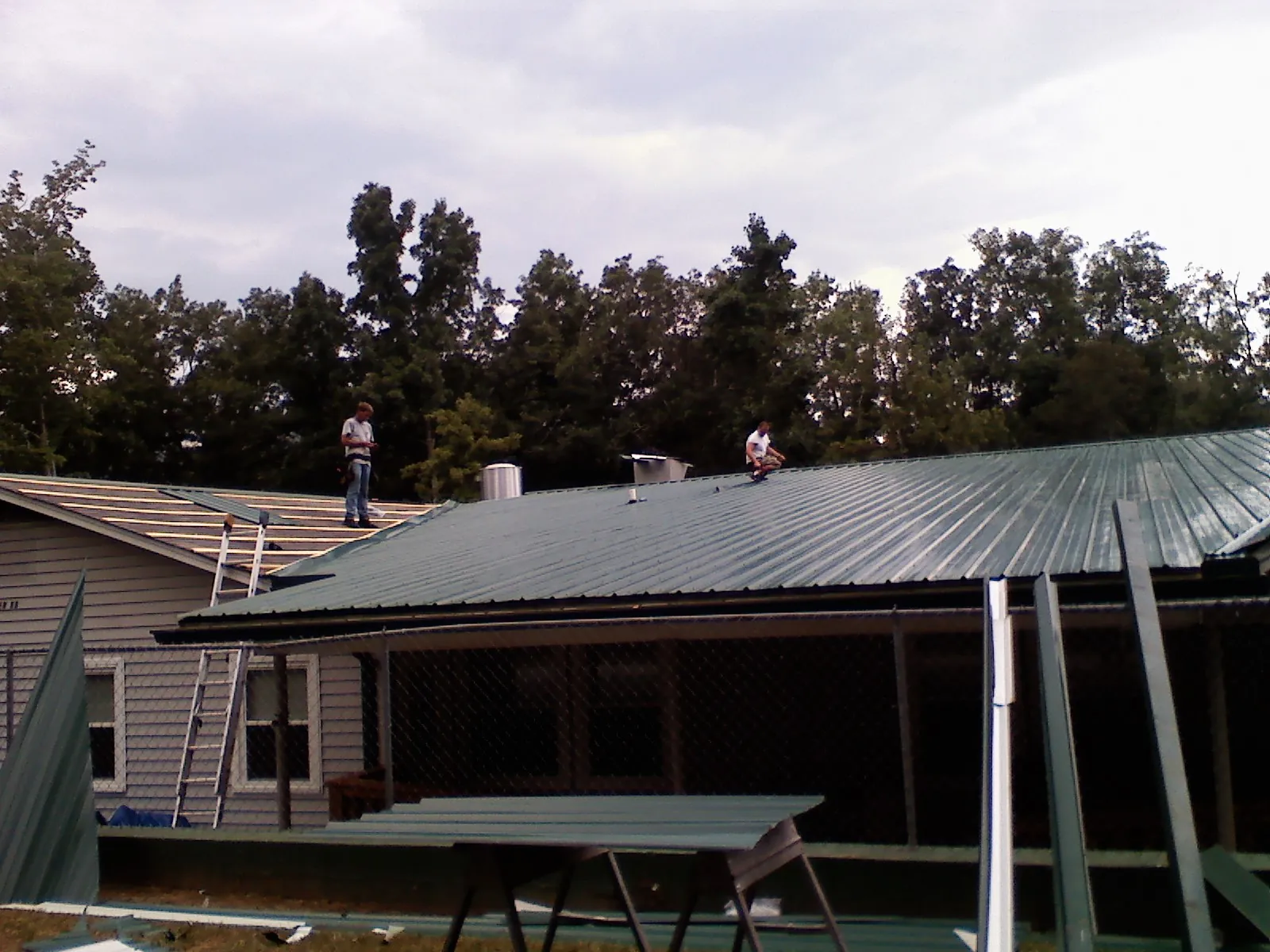 Two roofers installing green metal panels above a covered porch in Molalla
